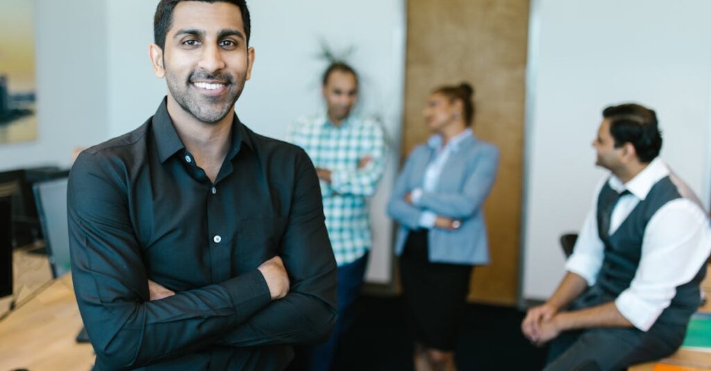 A confident young professional standing in a modern office with colleagues interacting in the background.